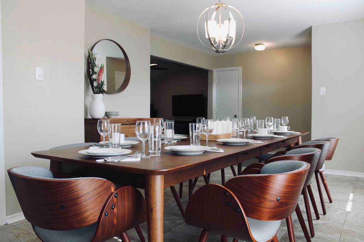 A modern dining area is showcased with a long wooden table, set for a meal with elegant dinnerware and glassware. Contemporary chairs are arranged around the table. Soft lighting is provided by a stylish chandelier overhead, enhancing the welcoming ambiance.