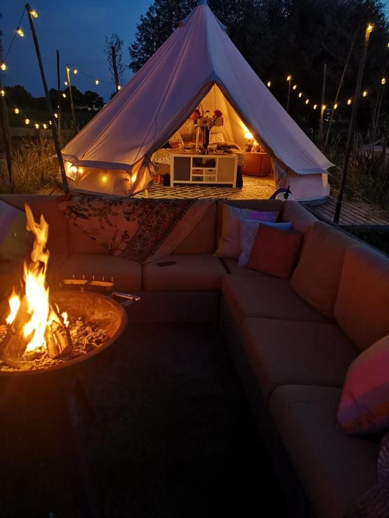 A cozy seating area is highlighted by a fire pit, surrounded by a circular lounge couch adorned with colorful cushions. In the background, the bell tent is illuminated by warm lights, creating an inviting atmosphere under the evening sky.