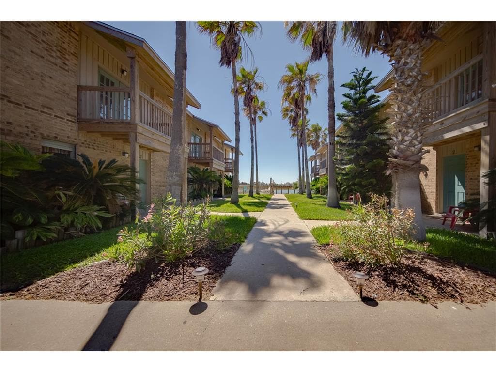 A well-maintained courtyard is framed by palm trees and lush greenery. A concrete pathway leads through the space, connecting the buildings. Bright sunlight illuminates the area, highlighting the vibrant plants flanking the walkway.