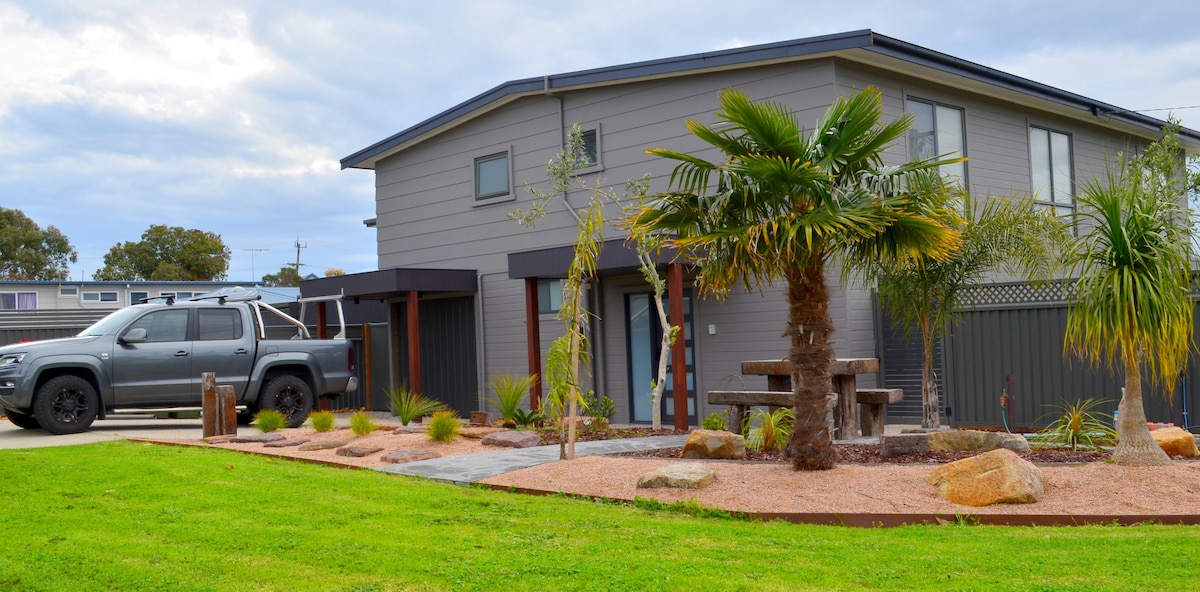 The exterior of the modern home is displayed, featuring a well-maintained garden with tropical plants and decorative stones. A picnic table is situated in the front yard, surrounded by a neatly manicured lawn. The exterior walls are painted in neutral tones under a slightly cloudy sky.