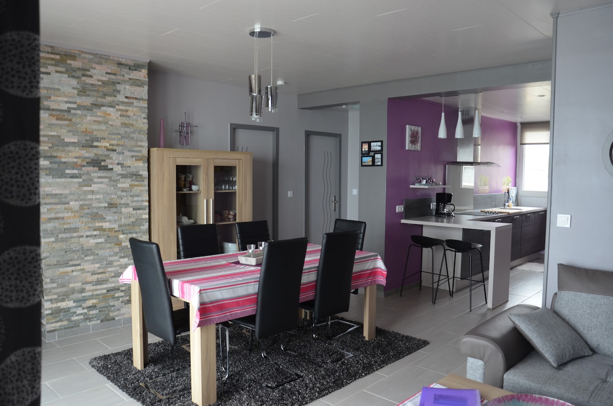 A spacious dining area features a rectangular table with black chairs, adorned with a colorful tablecloth. Light tones of the walls complement the modern kitchen, visible in the background, which includes sleek cabinetry and appliances. Natural light enters through large windows.