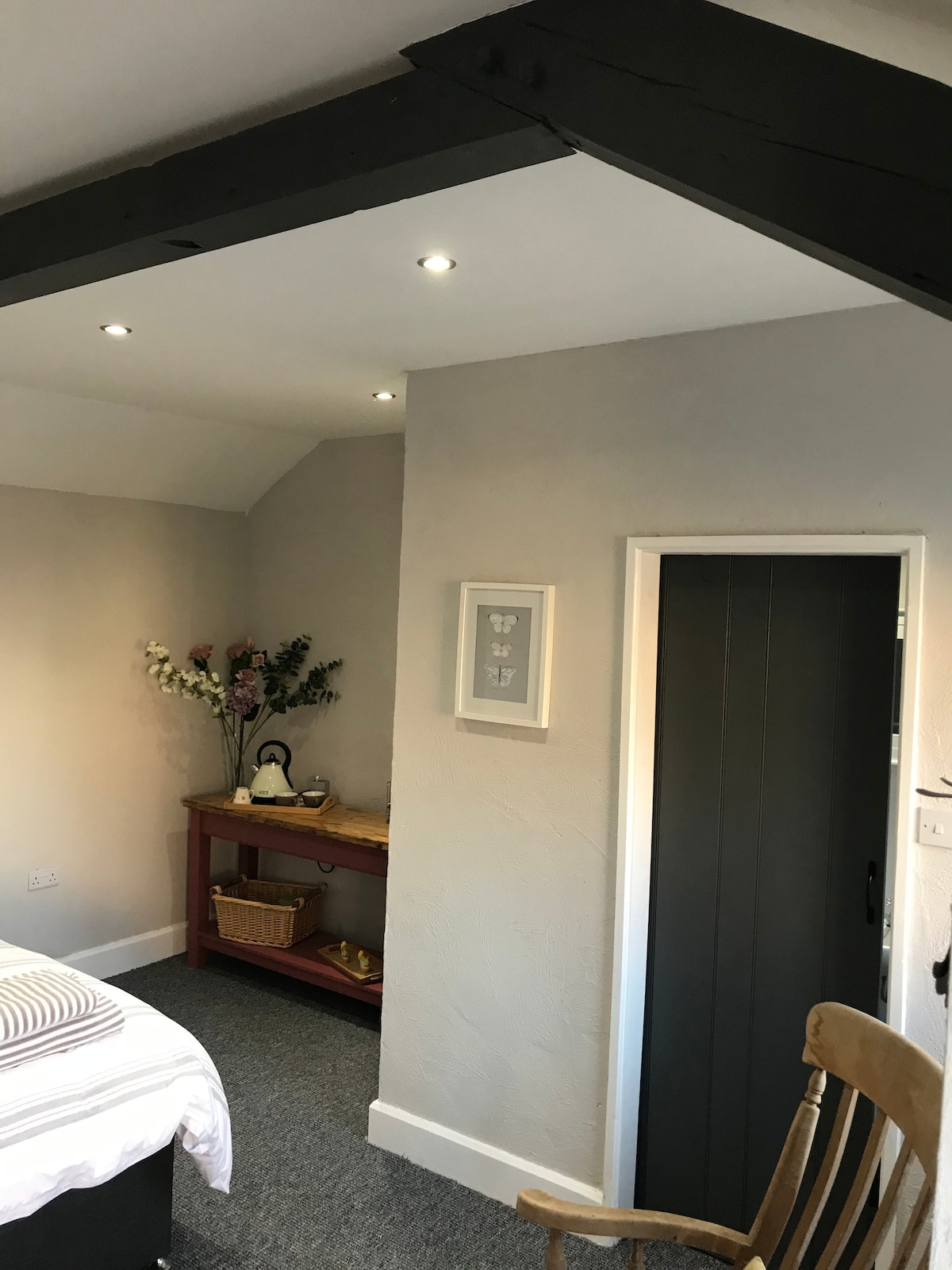A well-lit corner of the room features a wooden table with a woven basket and a small kettle setup. A framed art piece hangs on the wall, complemented by a vase of flowers. A dark door leads to the en-suite bathroom.