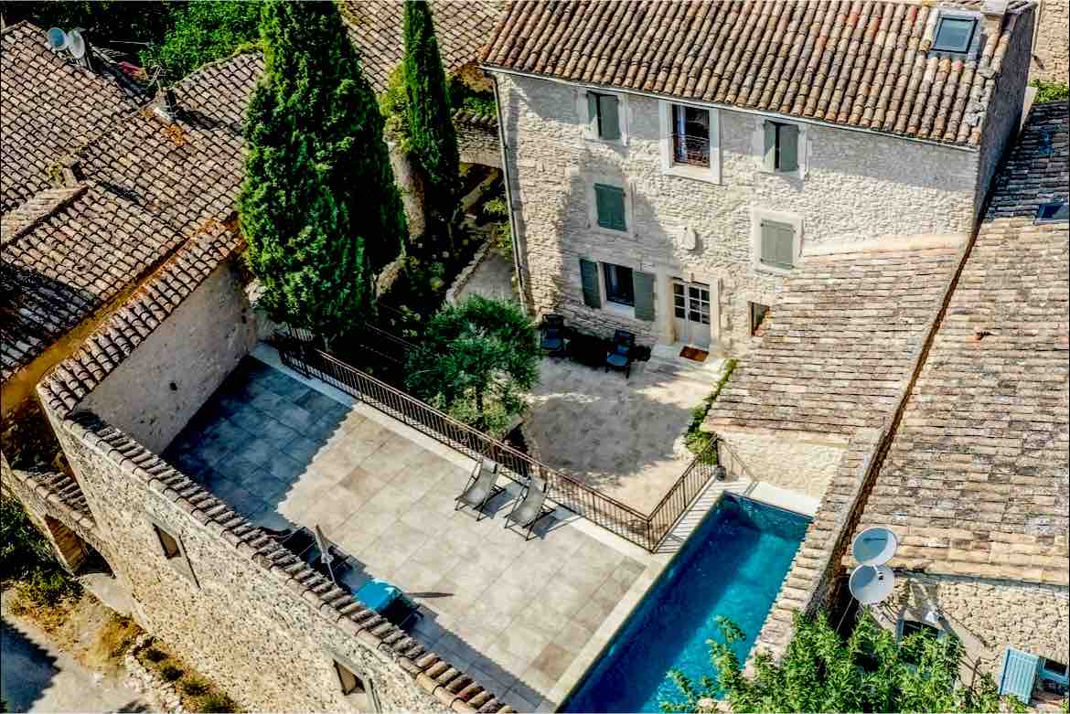 An aerial view showcases a charming stone building surrounded by lush greenery. A tranquil swimming pool is located in a spacious courtyard, bordered by a secure fence. Sun loungers are positioned around the pool area, and traditional rooftops can be seen nearby.
