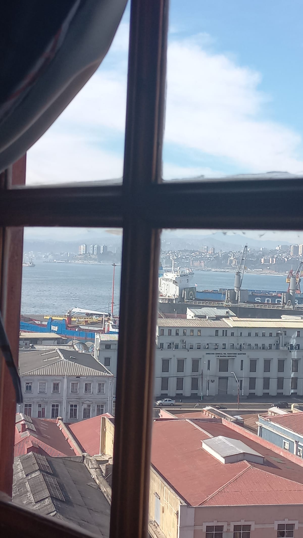 A view of the bay is seen through a window, framed by wooden panes. The scene captures boats docked on the water and the historic buildings lining the waterfront, with a backdrop of hills and cloudy skies.