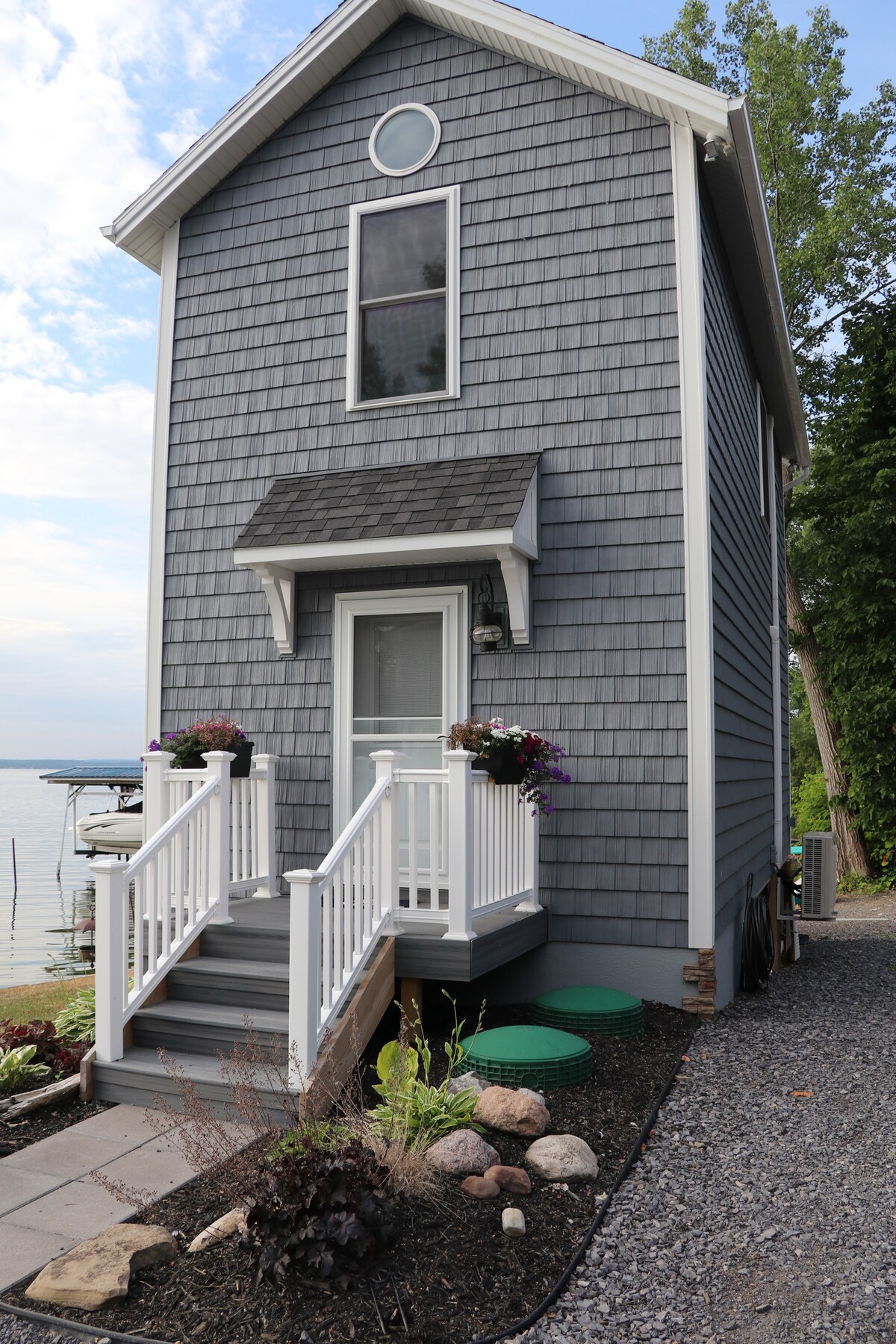 The exterior of a two-story bunkhouse is shown, featuring gray shingles and white trim. A welcoming entrance with a set of steps is flanked by potted flowers. The building overlooks a lake, with a smooth pathway lined with gravel and landscaping elements such as rocks and plants.