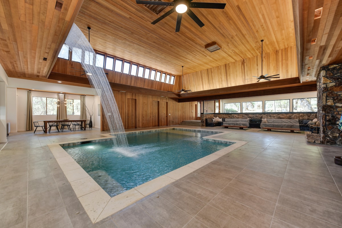 An indoor pool is centered in a spacious room, surrounded by natural wood paneling and large windows that invite abundant light. A water feature cascades gently into the pool, while a lounge area with comfortable seating is positioned nearby, creating a relaxing environment.