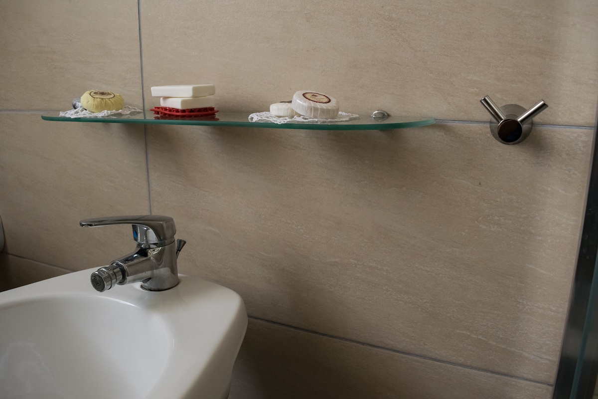 A clean bathroom corner is presented, featuring a sleek sink with a modern faucet. Above the sink, a glass shelf displays various toiletries, including soaps and a small decorative item. The neutral-toned tiles add to the overall simplicity of the space.