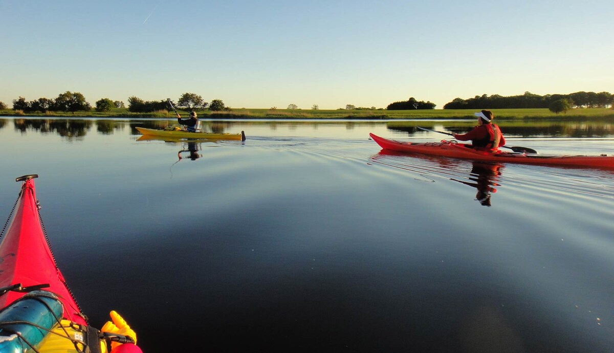 Two kayakers navigate a calm waterway, surrounded by lush green landscape. The glassy surface of the water reflects the clear blue sky, while gentle ripples trail behind the kayaks. The scene captures a moment of tranquility and connection with nature.