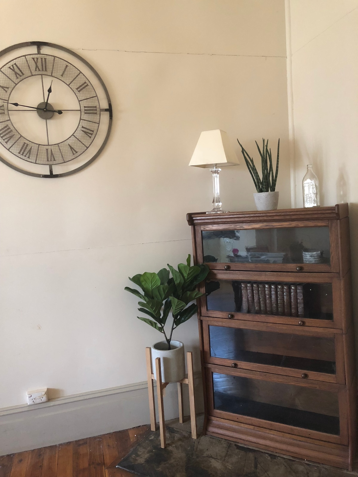 A corner of the room features a vintage wooden cabinet with glass doors, displaying books and decorative items. Above, a large wall clock with Roman numerals is mounted. A small lamp and a potted plant sit atop the cabinet, while a tall planter holds a leafy green plant.