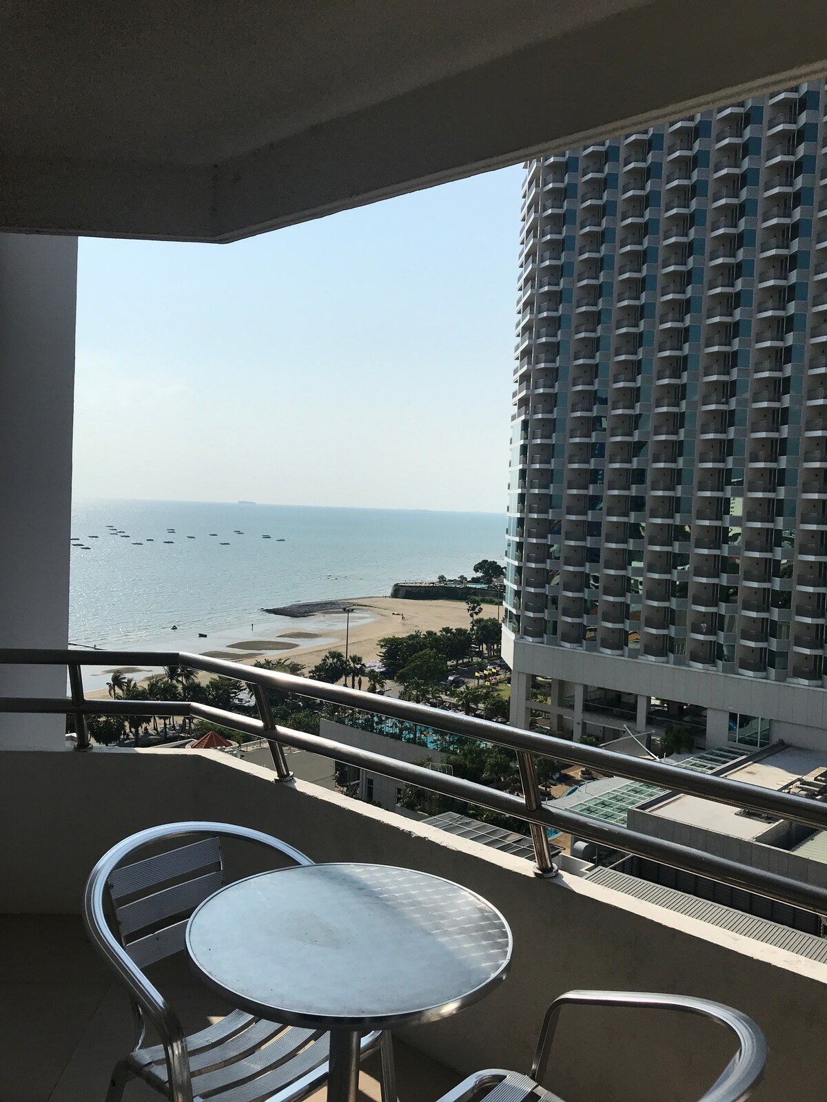 A balcony view displays shimmering ocean waters and a sandy beach. In the background, a modern high-rise building rises alongside the coastline. A small, round table and two chairs are positioned for guests to enjoy the surroundings.