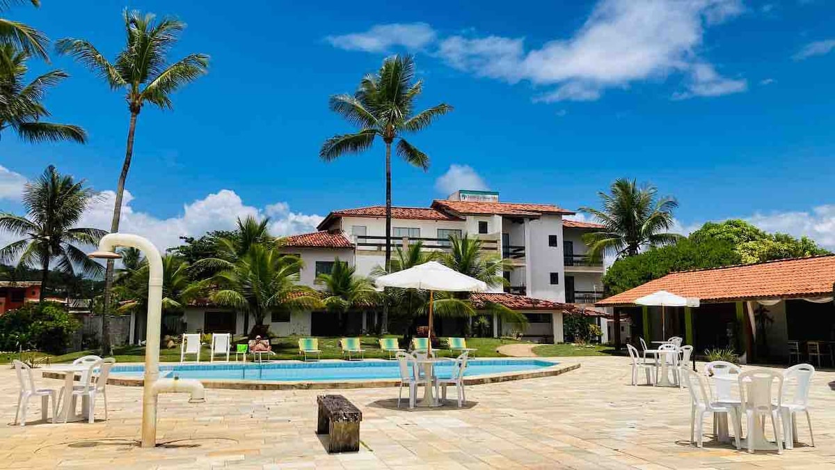 A sparkling pool is surrounded by lounge chairs and umbrellas, set against a backdrop of lush palm trees and a sunny sky. The hotel’s modern structure is visible in the distance, featuring multiple levels and a tile roof. A relaxed atmosphere is suggested by the open space.