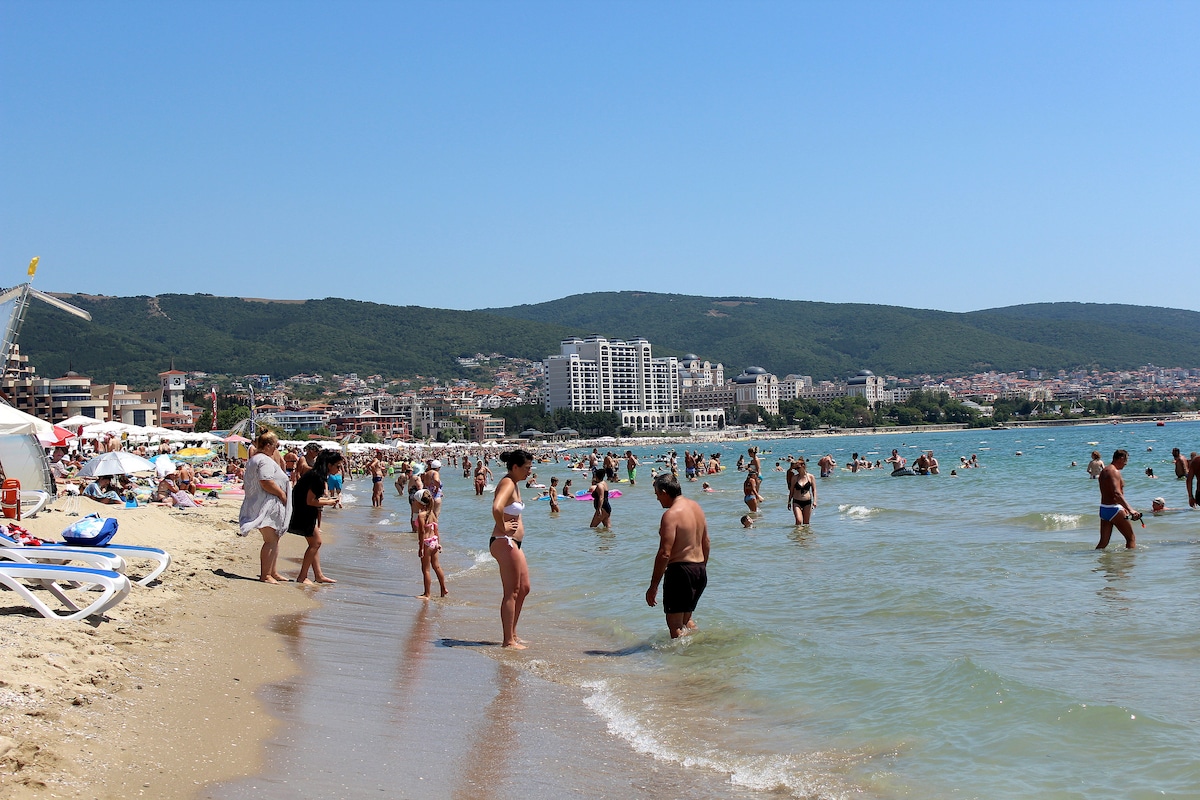 The image depicts a busy beach scene under clear blue skies. People are seen enjoying the water, with some walking along the shore. Beach umbrellas and lounge chairs are placed on the sand, while buildings are visible in the background, overlooking the beach area.