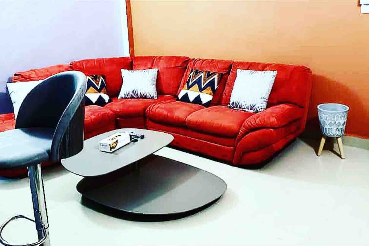A spacious living area is furnished with a red sectional sofa, featuring decorative cushions with geometric patterns. A sleek, oval coffee table sits in front, accompanied by a modern bar stool. A small decorative basket adds a touch of style to the corner.