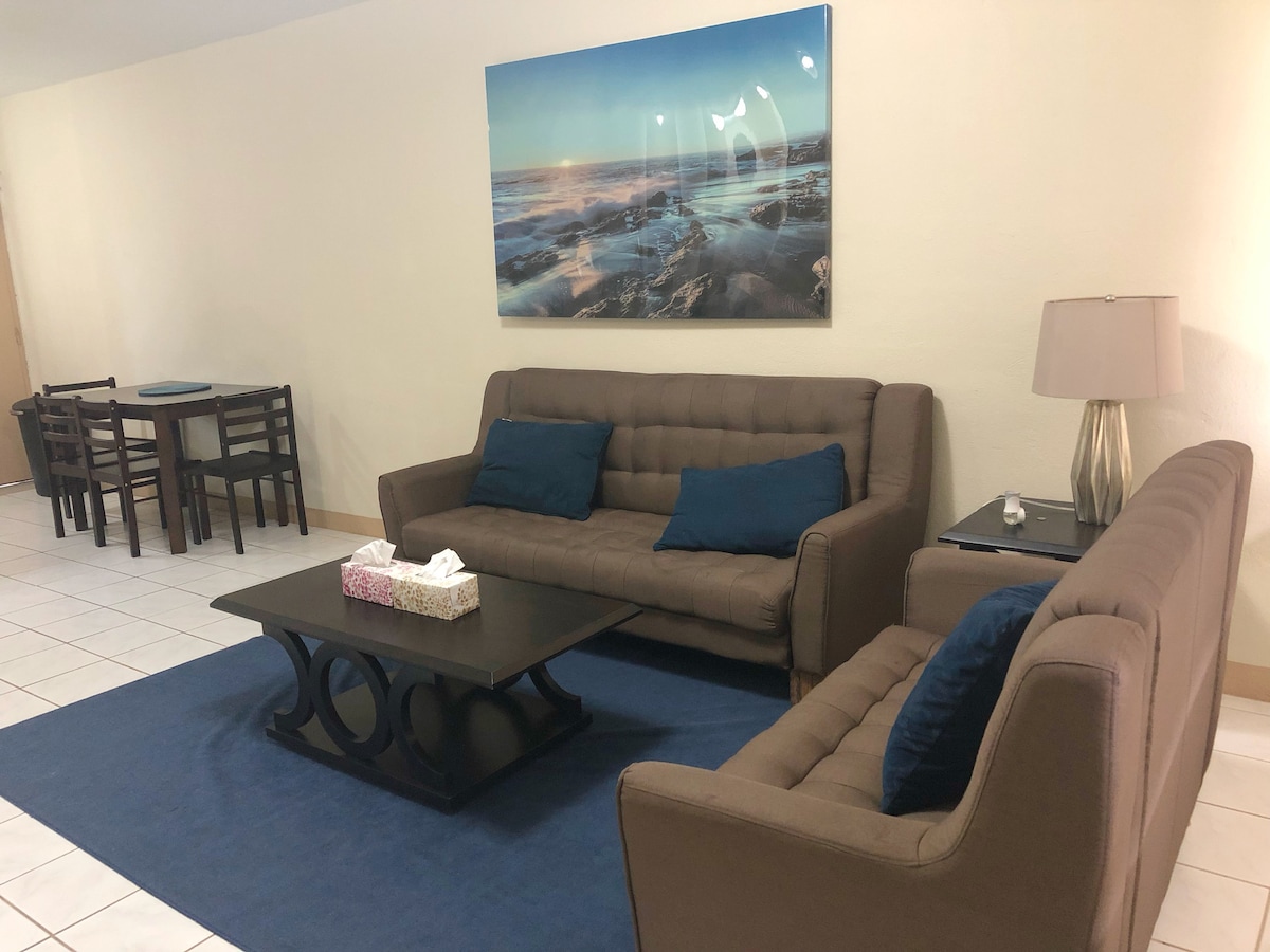 A spacious living area features two sofas adorned with blue cushions, positioned around a central coffee table. A dining table with four chairs is visible in the background. A large wall-mounted artwork depicts a serene ocean scene, contributing to the calm ambiance.