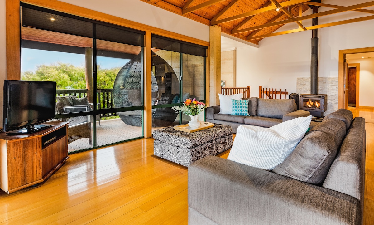 An open-plan living area features a comfortable seating arrangement with a large sofa, a coffee table, and a flat-screen TV. Natural light pours in through expansive glass doors that lead to a deck, showcasing a cozy fireplace and warm wooden accents.