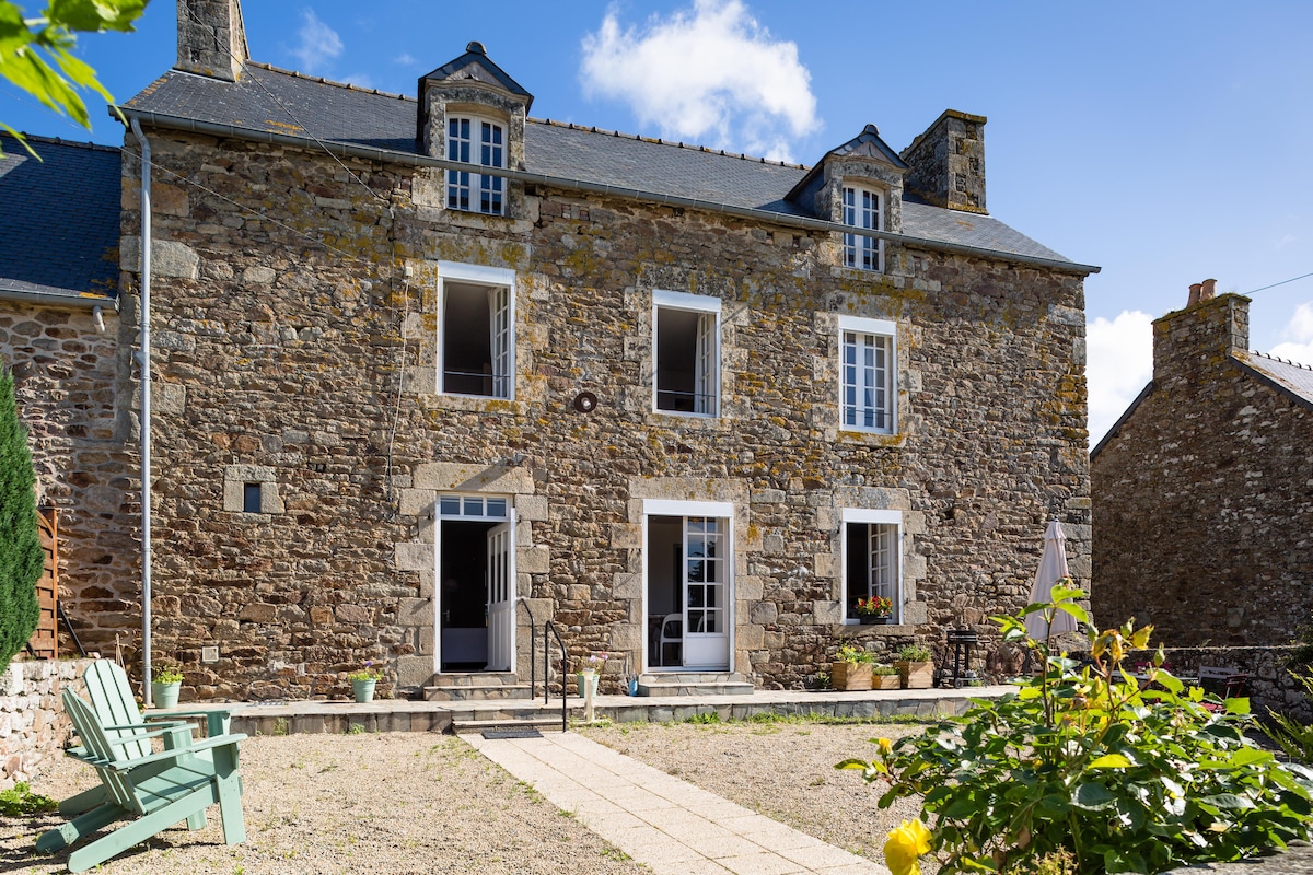 The exterior of a stone house is shown, highlighting three upper windows and a central glass door framed by flower pots. A square pathway leads to the entrance, with seating areas visible on either side. The surrounding area features greenery and a clear blue sky.