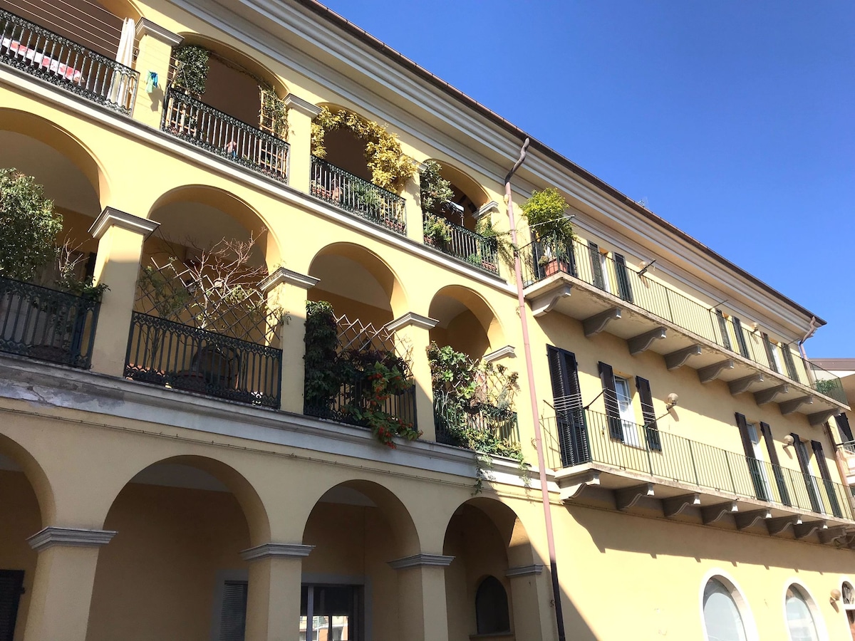 An historic building is displayed with multiple covered balconies adorned with plants and vibrant greenery. Arched openings create a rhythm across the façade, while a clear blue sky provides a contrasting backdrop, enhancing the architectural details.