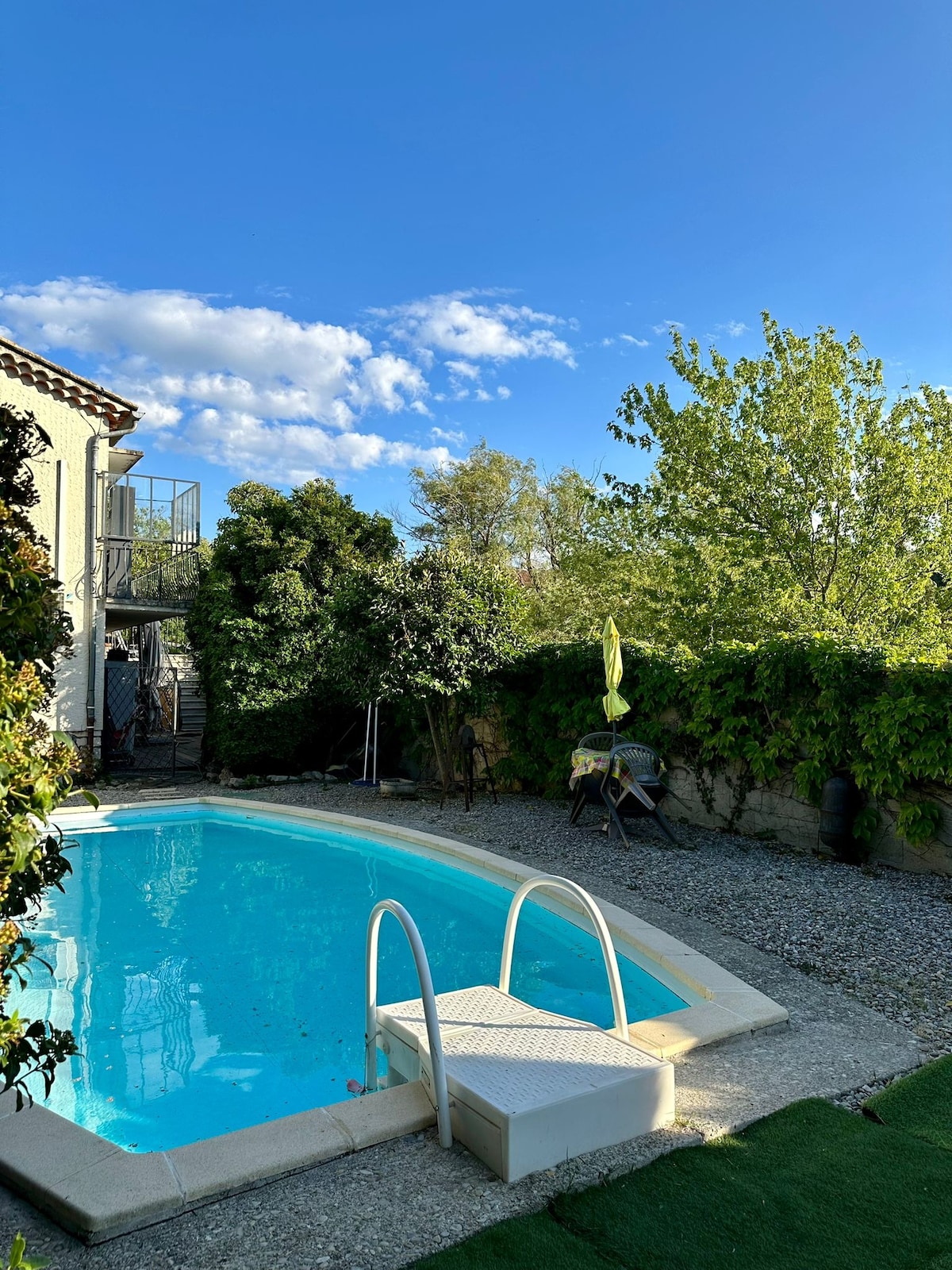 A serene private pool is featured, surrounded by lush greenery and a tranquil garden. A small ladder leads into the clear blue water, reflecting the clear sky above. A lounge area with a chair is visible, offering a peaceful spot for relaxation.