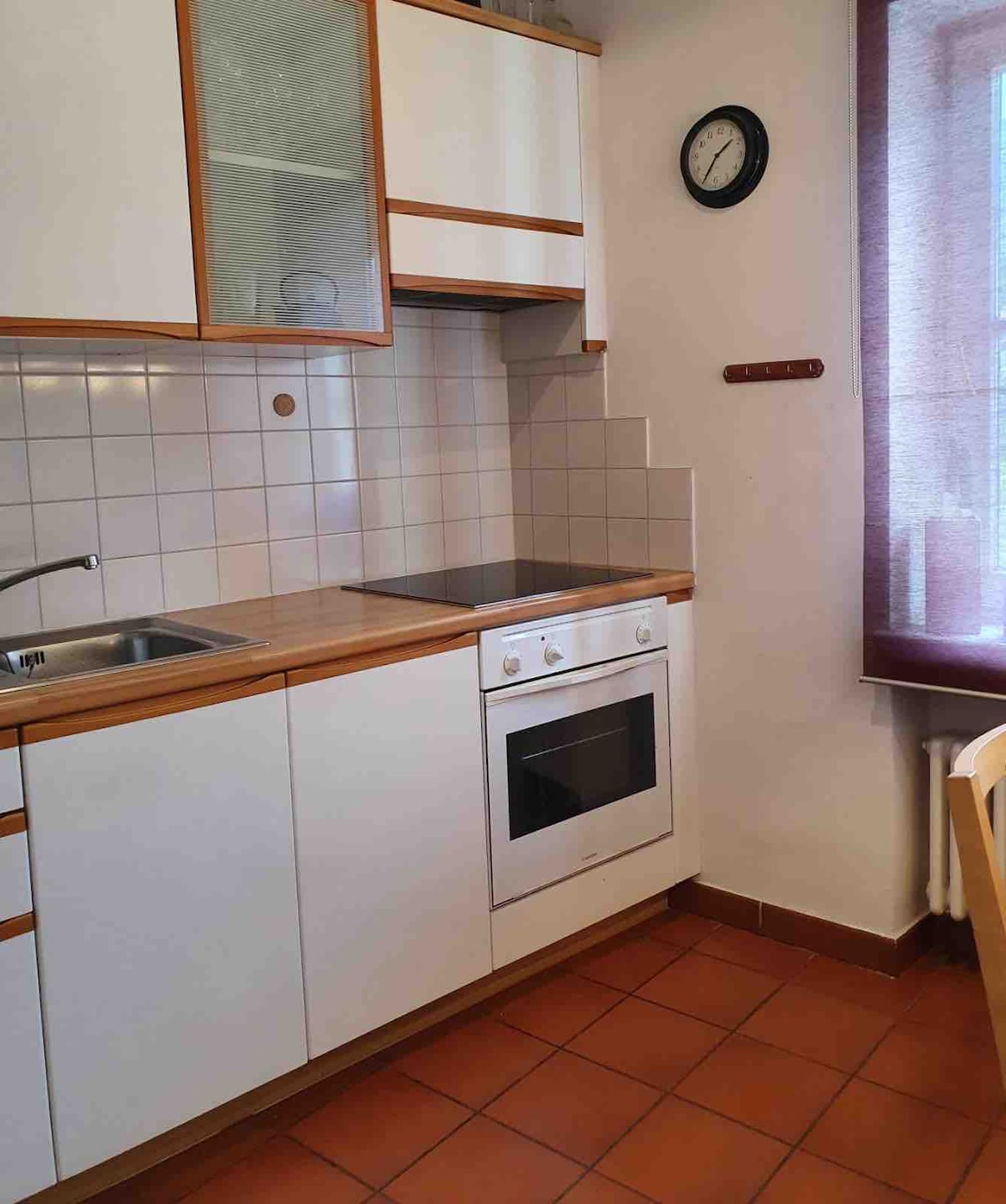 A compact kitchen area showcases sleek white cabinetry accented with wooden trim. Stainless steel sinks are positioned next to a built-in oven and stove. Natural light filters through a nearby window, illuminating the tiled floor and creating a functional cooking space.