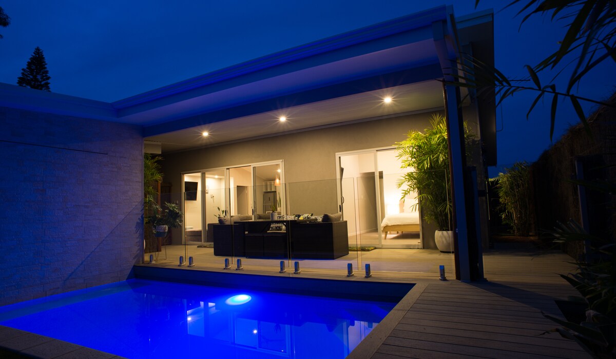 A modern villa is illuminated at night, showcasing a private plunge pool with glowing blue water. Large glass doors provide a view of the stylish interior, where a comfortable seating area is visible. Tropical plants frame the entrance, enhancing the serene outdoor space.
