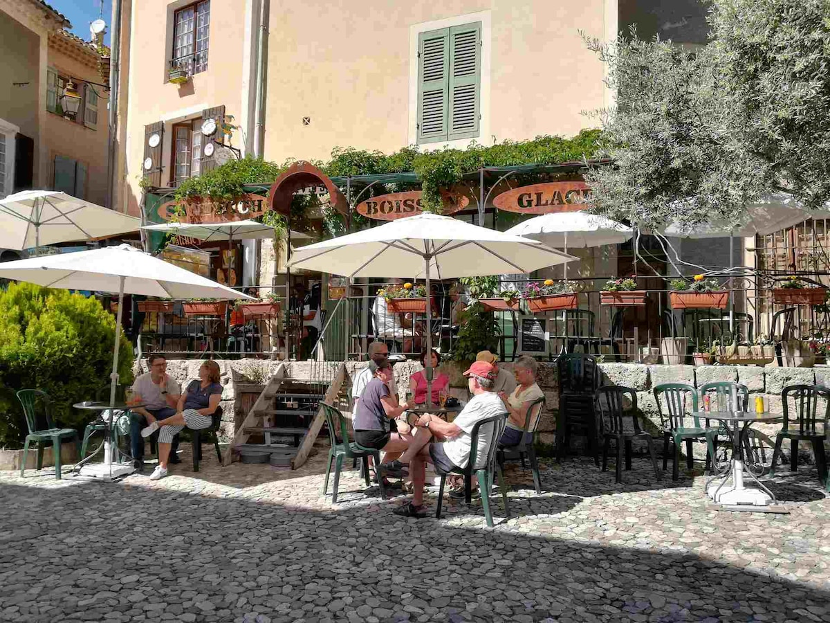 An outdoor café setting is depicted with several patrons seated at green metal chairs around round tables. Large umbrellas provide shade over the tables, while planters with greenery are positioned against the building backdrop, enhancing the cafe's inviting atmosphere.
