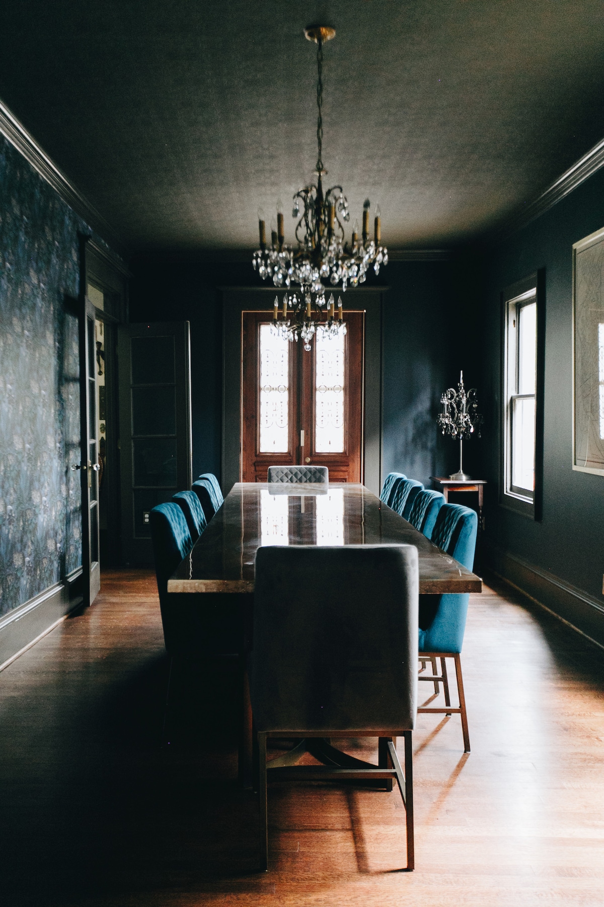 A long dining table is surrounded by upholstered chairs in a deep blue hue. A crystal chandelier hangs from the ceiling, adding elegance. The walls are adorned with a subtle floral wallpaper, and a large window allows soft light to enter the room.