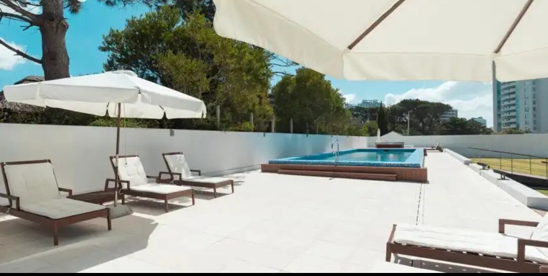 A sunlit outdoor area features a heated pool surrounded by a spacious terrace. Sun loungers are positioned under large umbrellas, providing shaded spots for relaxation. Lush trees frame the setting, offering a serene environment for guests enjoying the poolside facilities.