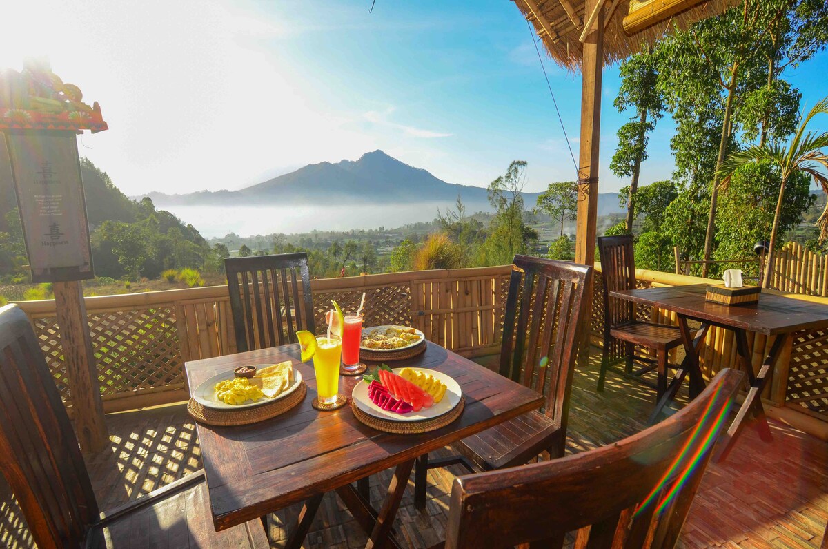 A wooden dining area is shown outdoors, featuring a table set with colorful drinks and an array of fresh fruit. Lush greenery surrounds the space, with views of distant mountains under a bright sky. The morning light enhances the serene setting.