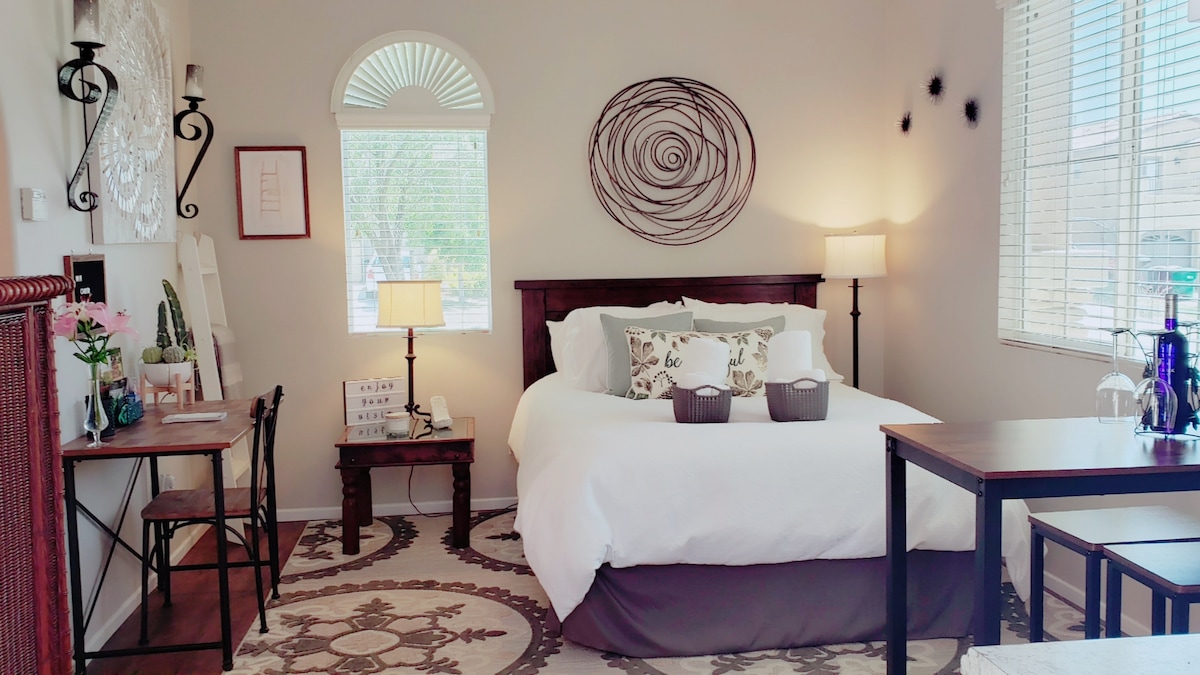 A well-appointed room features a queen bed dressed in soft linens, complemented by decorative pillows. A charming artwork hangs above the headboard. Natural light enters through a nearby window, illuminating a small dining table and two chairs, enhancing the cozy ambiance.