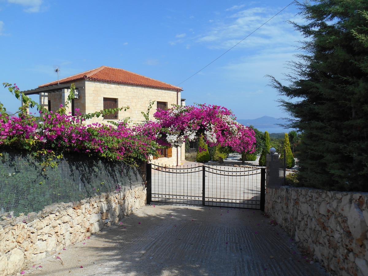 A stone villa is framed by vibrant pink bougainvillea, creating a welcoming entrance. The gated driveway leads up to the property, flanked by well-maintained greenery. Expansive views of the countryside and mountains are visible in the distance against a clear blue sky.