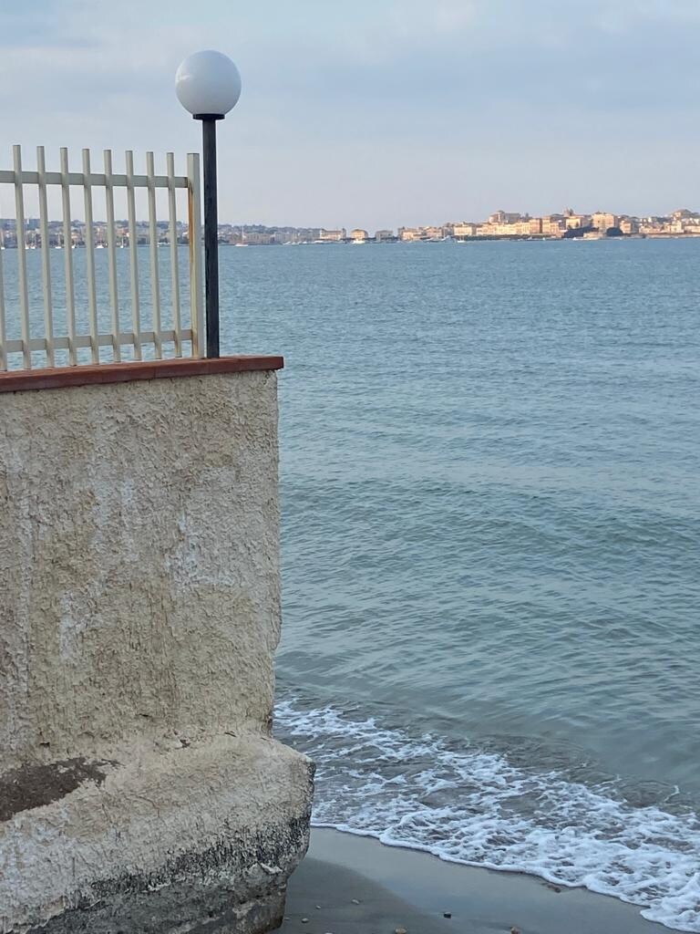 The image captures a coastal view featuring gentle waves lapping against the shore. A light pole is positioned near the water's edge, while a low wall made of stone and concrete stands beside it. In the distance, a shoreline can be seen with buildings along the horizon.