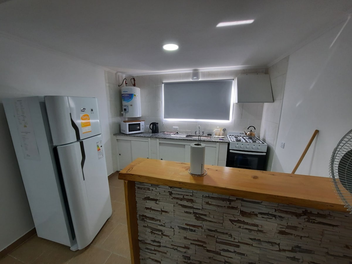 A modern kitchen is displayed with light-colored cabinetry and a spacious countertop. Essential appliances, including a refrigerator, stove, and microwave, are neatly arranged. A window allows natural light to brighten the space, while a ceiling light fixture provides additional illumination.