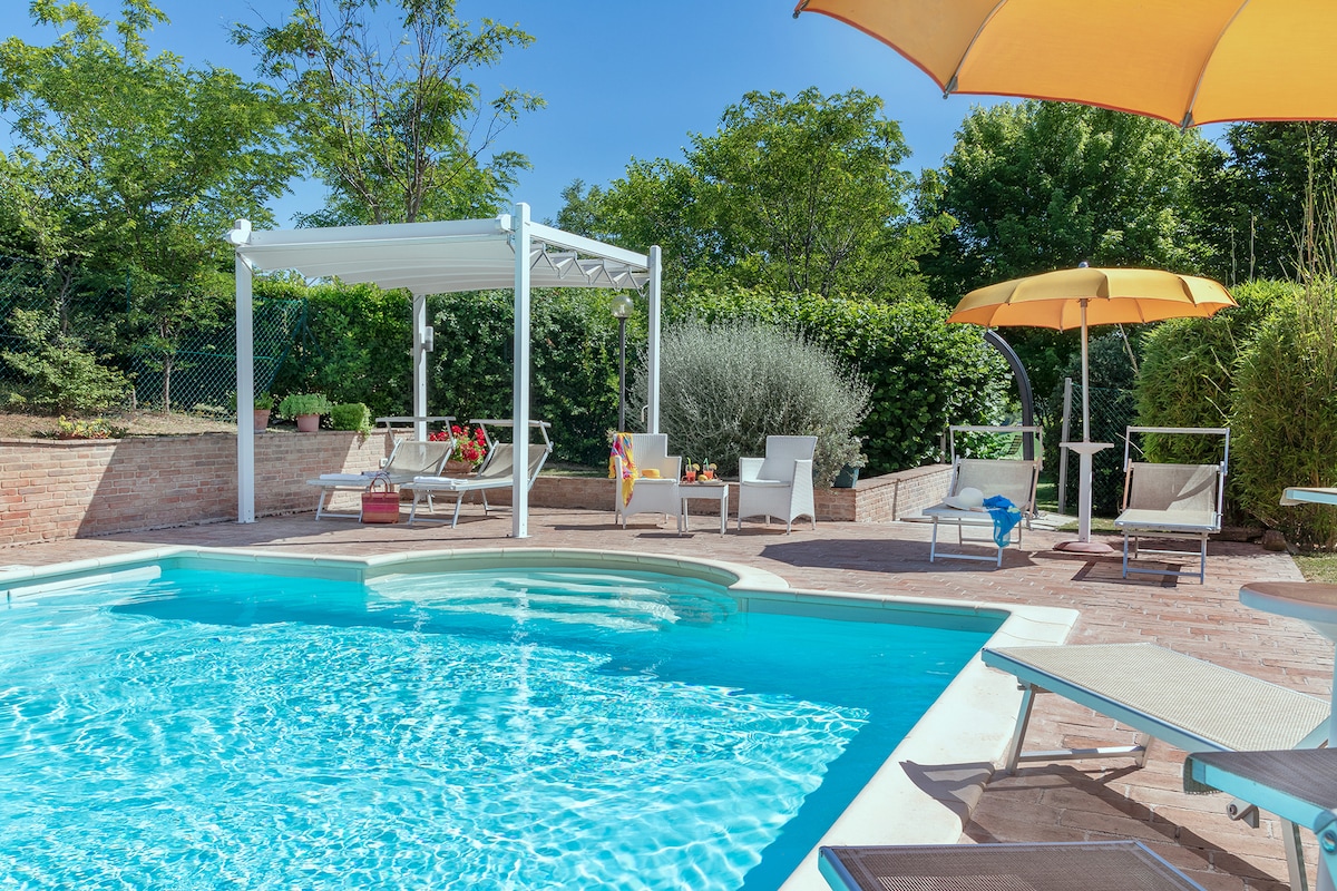 A serene outdoor area features a rectangular salt-water pool, accompanied by a white gazebo and sun loungers. A large shaded umbrella offers protection from the sun, and lush greenery surrounds the space, providing a tranquil ambiance for relaxation.