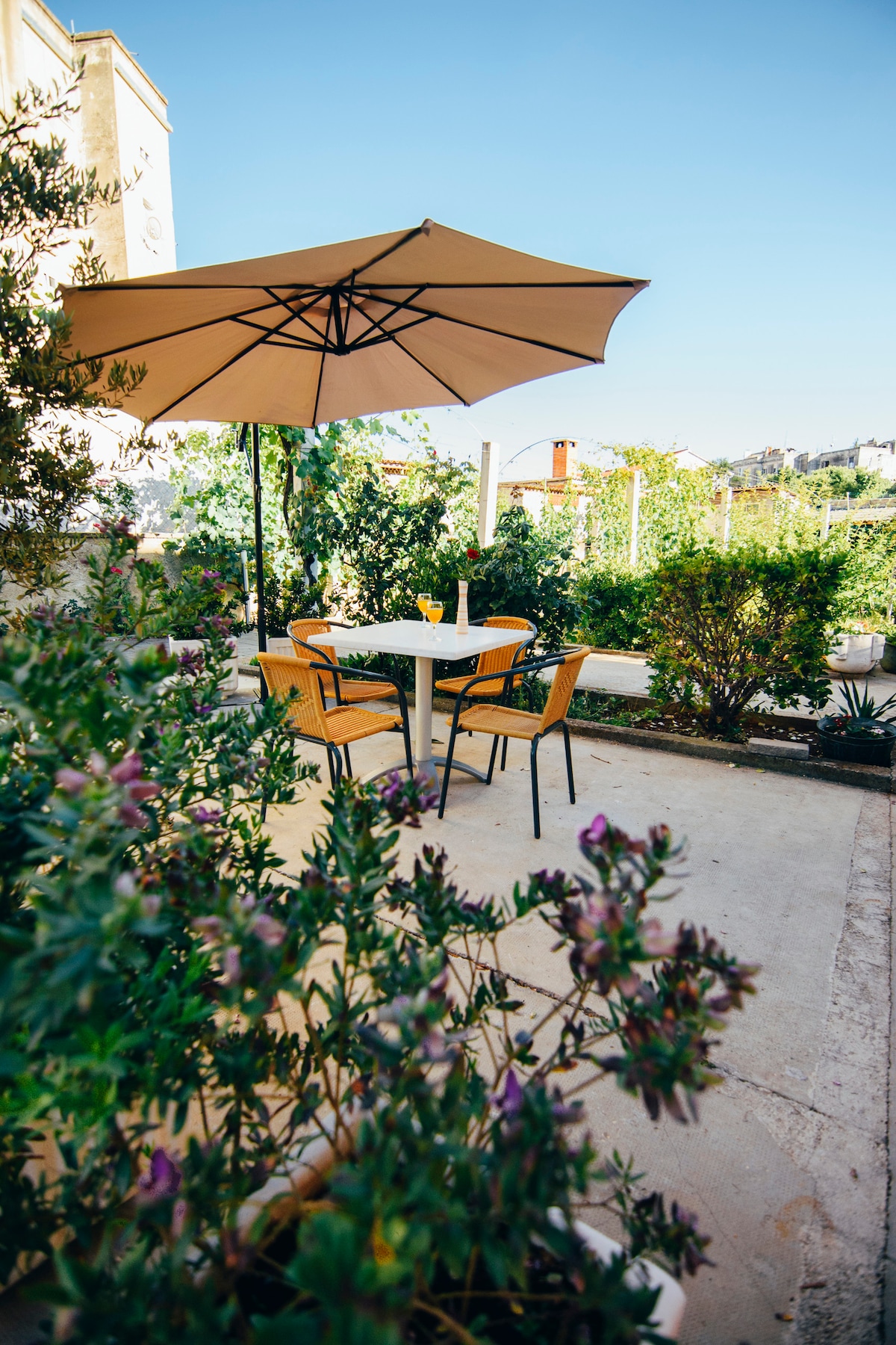 An outdoor seating area is presented with a round table and four chairs, shaded by a large umbrella. Surrounding greenery, including flowering plants, creates a relaxed environment. The concrete floor provides a neutral backdrop, enhancing the space's inviting feel.