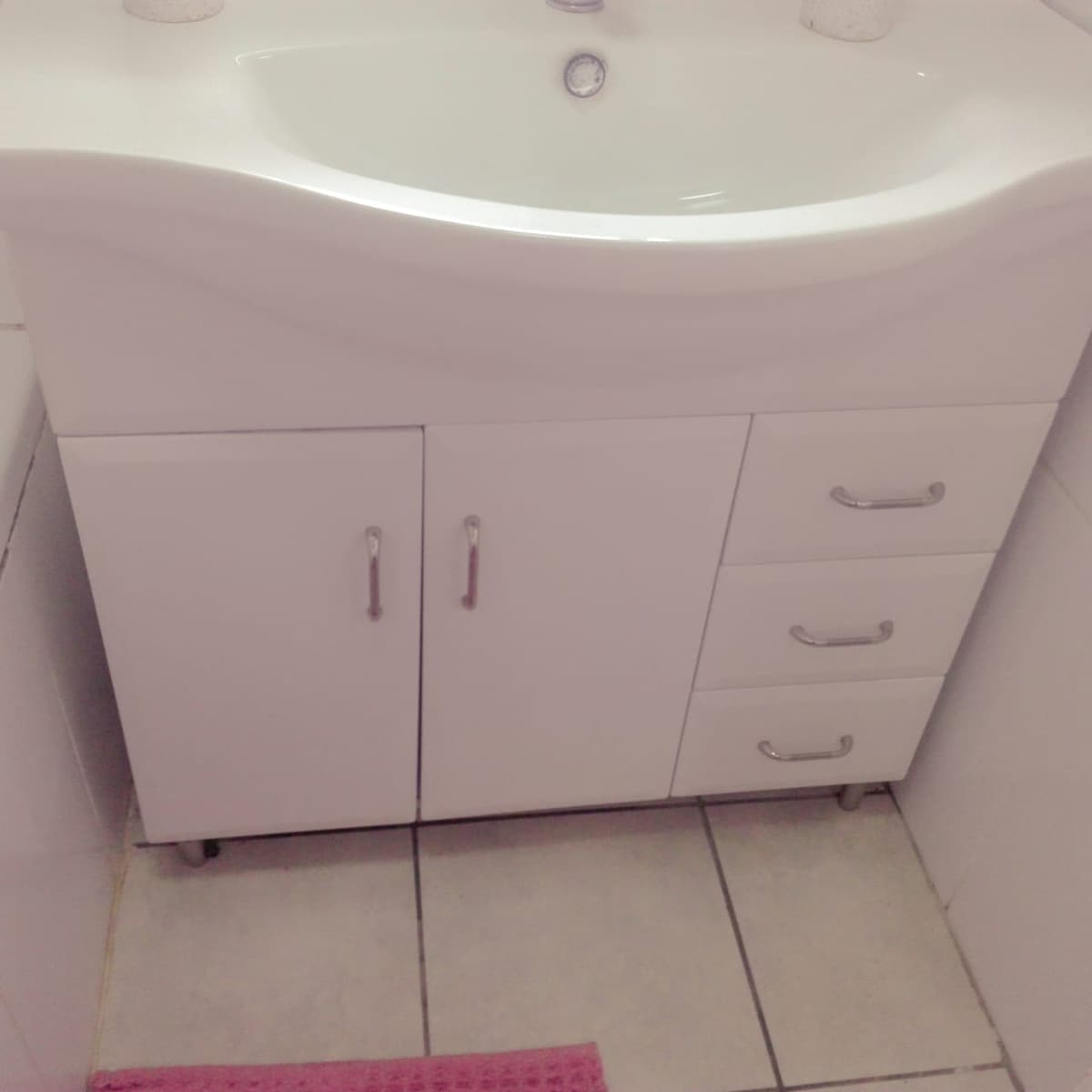 A modern bathroom sink unit is featured, composed of a smooth, white surface and sleek silver handles. Below, a cabinet with two doors and three drawers provides storage. The floor is covered with light tiles, and a pink mat adds a touch of color nearby.