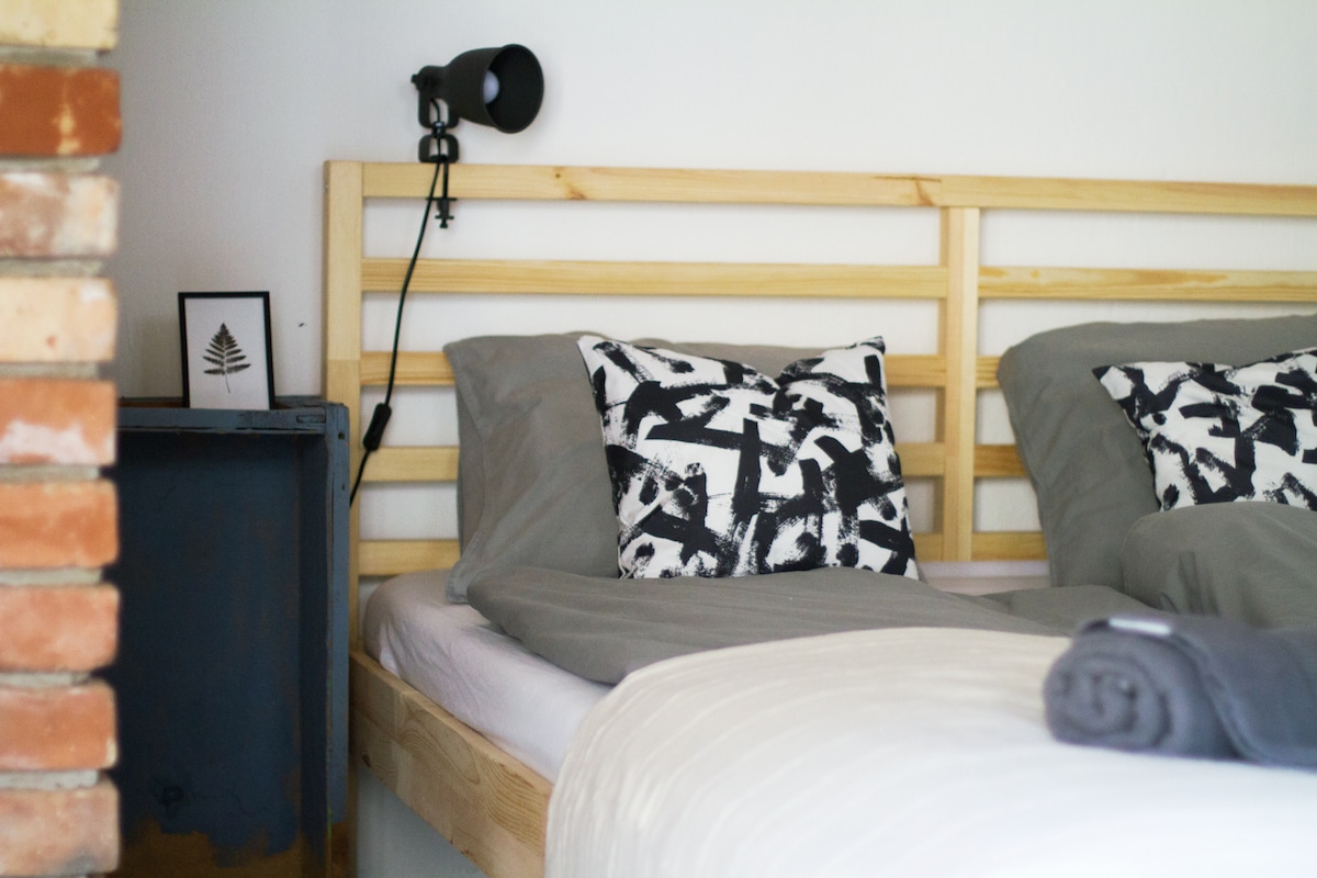 A cozy sleeping area features a wooden bed frame with two beds dressed in gray linens. Each bed is adorned with decorative pillows showcasing black and white patterns. A bedside table holds a minimalist framed artwork, while a wall lamp provides soft lighting.