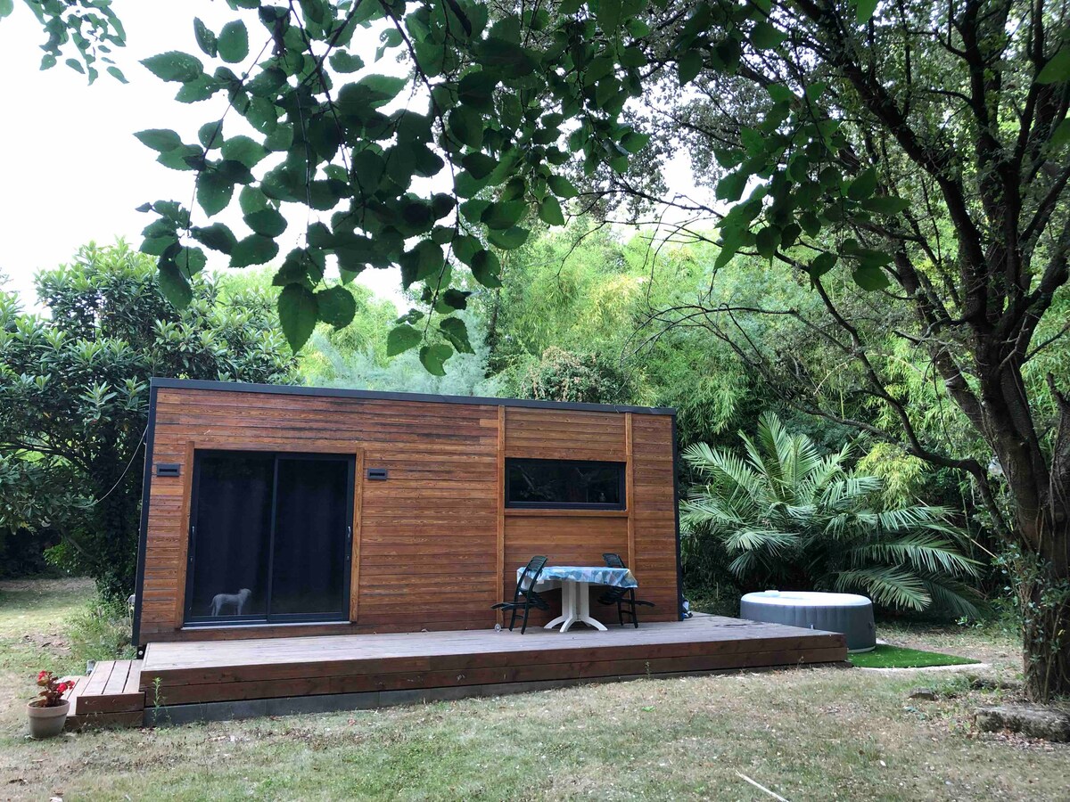 A wooden tiny house is set amidst lush greenery, featuring a spacious deck with a table and two chairs. A hot tub can be seen on the side, surrounded by tranquil landscaping, providing a serene outdoor space.