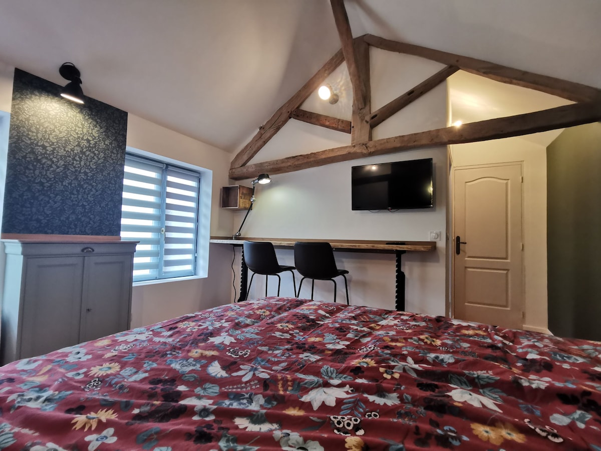 A cozy interior features exposed wooden beams and a floral-patterned bedspread on a spacious bed. A small workspace with two chairs and a wall-mounted television is visible, accompanied by a window with horizontal blinds that allows natural light to fill the room.