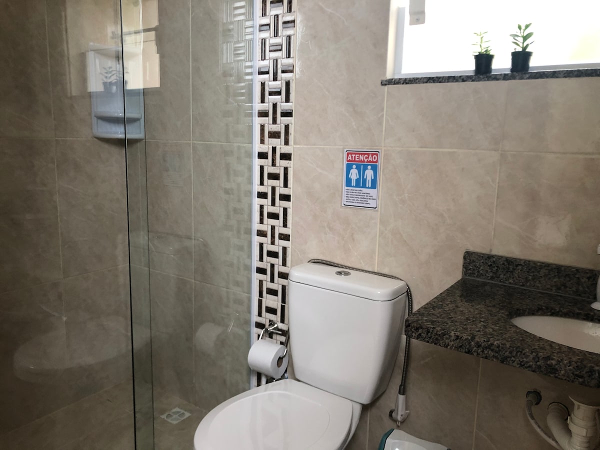 The bathroom is equipped with a modern toilet and a glass shower enclosure. A granite countertop holds a sink and essential toiletries are visibly organized. Green plants sit atop a nearby shelf, adding a touch of nature to the ambiance.
