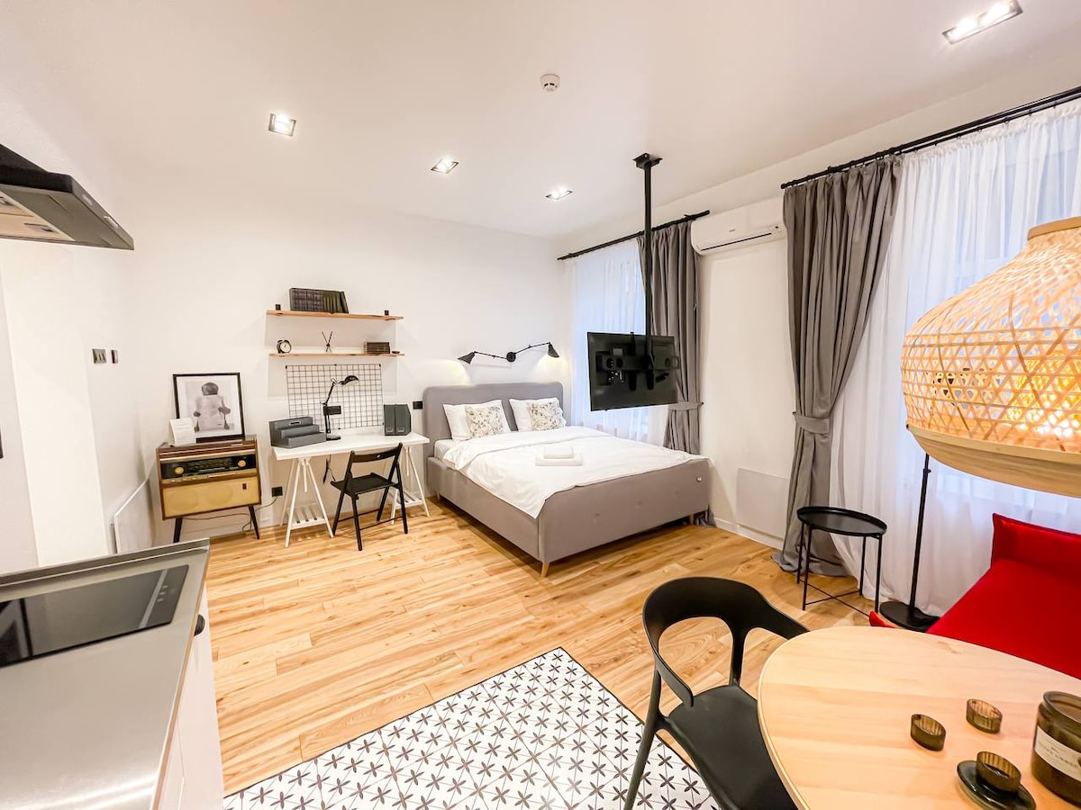 A stylish studio space features a spacious bed set against light walls. A wooden floor complements modern furniture, including a round dining table and black chairs. Large windows allow natural light, while a unique pendant lamp and wall-mounted TV enhance the atmosphere.
