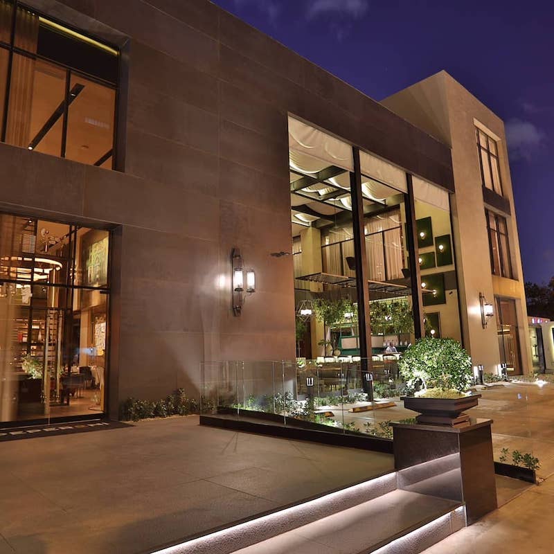 The building features a contemporary facade with large glass windows, allowing for a view of the illuminated interior. Soft outdoor lighting highlights the entrance, complemented by manicured greenery along the pathway. The modern design conveys elegance and sophistication.