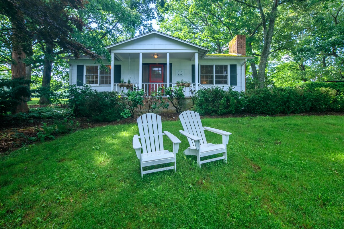 A charming cottage exterior is surrounded by lush greenery and tall trees. Two white Adirondack chairs face each other on a well-maintained lawn, inviting relaxation. The front porch features a welcoming red door, framed by evenly spaced windows providing views of the tranquil landscape.