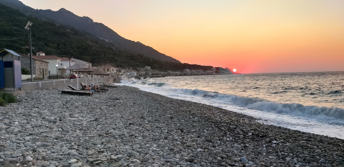 A serene beach scene is displayed, featuring a rocky shoreline with gentle waves lapping at the pebbles. The sunset casts warm hues across the sky and reflects on the water, while distant hills and scattered buildings are visible along the coast.