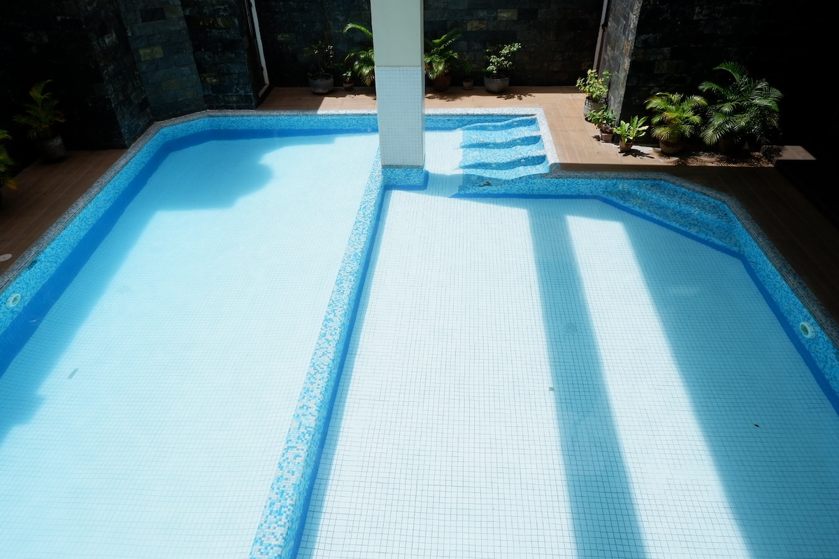 A refreshing outdoor swimming pool is featured, with clear blue water inviting relaxation. Surrounding the pool are potted plants, adding a touch of greenery to the space. Sunlight casts soft shadows on the tiled surface, enhancing the tranquil ambiance.