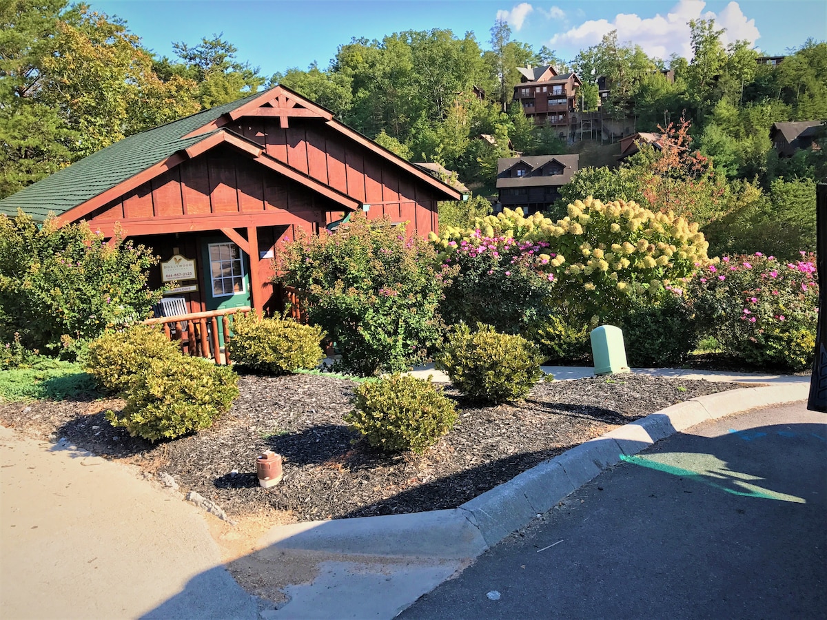 A charming red cabin is nestled among greenery, surrounded by blooming bushes and trees. The setting provides a peaceful atmosphere, with a gravel pathway leading toward the entrance. Nearby, additional cabins can be seen perched in the distance, enhancing the woodland feel.