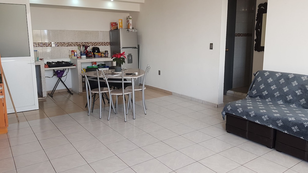 An open living space features a dining table surrounded by six chairs, with a kitchen visible in the background. A gray sofa is positioned against one wall. The tiled floor reflects natural light, creating a bright and spacious atmosphere.