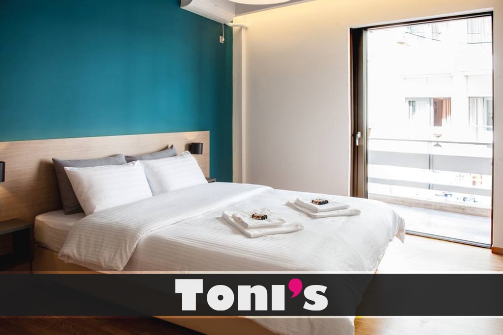 A well-organized bedroom is featured, showcasing a spacious bed with neatly arranged white linens and towels. A striking blue accent wall enhances the modern design, while natural light enters through a large window, which opens to a balcony view.