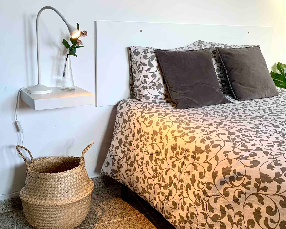 A cozy sleeping area features a spacious bed with a patterned duvet and two plush pillows. A bedside table with a reading lamp and a small vase of flowers is positioned beside the bed. A woven storage basket is placed on the floor.