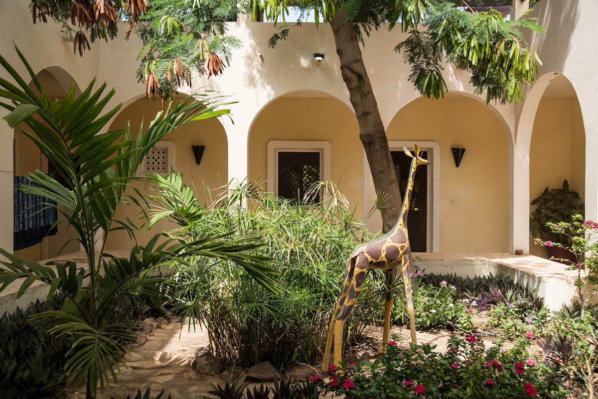 A vibrant courtyard is surrounded by soft yellow walls and arched doorways. Lush greenery and colorful flowers create a serene environment. A life-sized sculpted giraffe adds a whimsical touch to the garden, complemented by the shade of a large tree.