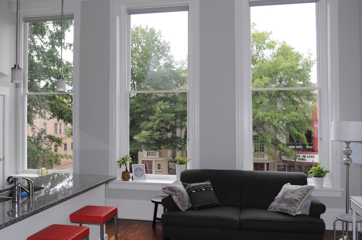 Tall windows frame a vibrant view of the neighborhood outside, letting in abundant natural light. A cozy seating area features a dark sofa adorned with decorative pillows. Behind, a modern kitchen bar with red stools and a glossy countertop is visible.